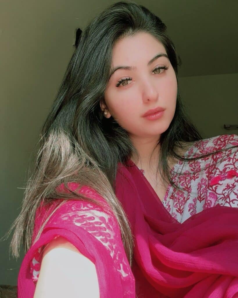 A woman with long dark hair wearing a pink and white patterned outfit and a bright pink scarf looks into the camera. Sunlight highlights her face and hair, with a neutral indoor background behind her.