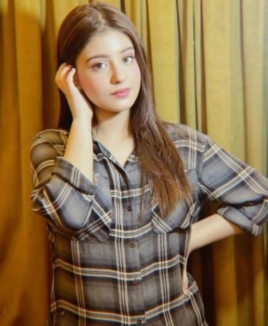 A young woman with long, straight brown hair stands indoors in front of brown curtains. She is wearing a gray plaid shirt, with one hand touching her hair and the other resting on her hip, looking at the camera.