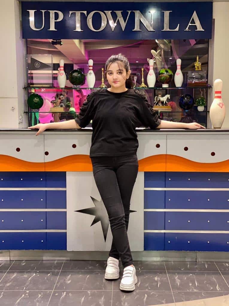 A young woman in black clothes and white sneakers stands in front of a bowling counter with bowling pins and balls, and a sign reading UPTOWN LA in the background.