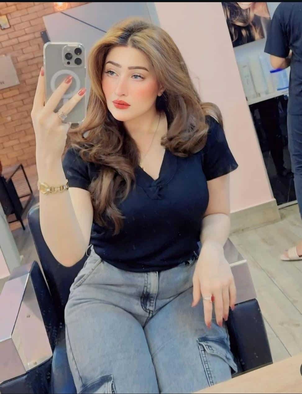 A woman with long, wavy light brown hair sits in a salon chair, wearing a black top and gray jeans, taking a mirror selfie with her phone. She has light makeup and red lipstick. The salon background is visible.