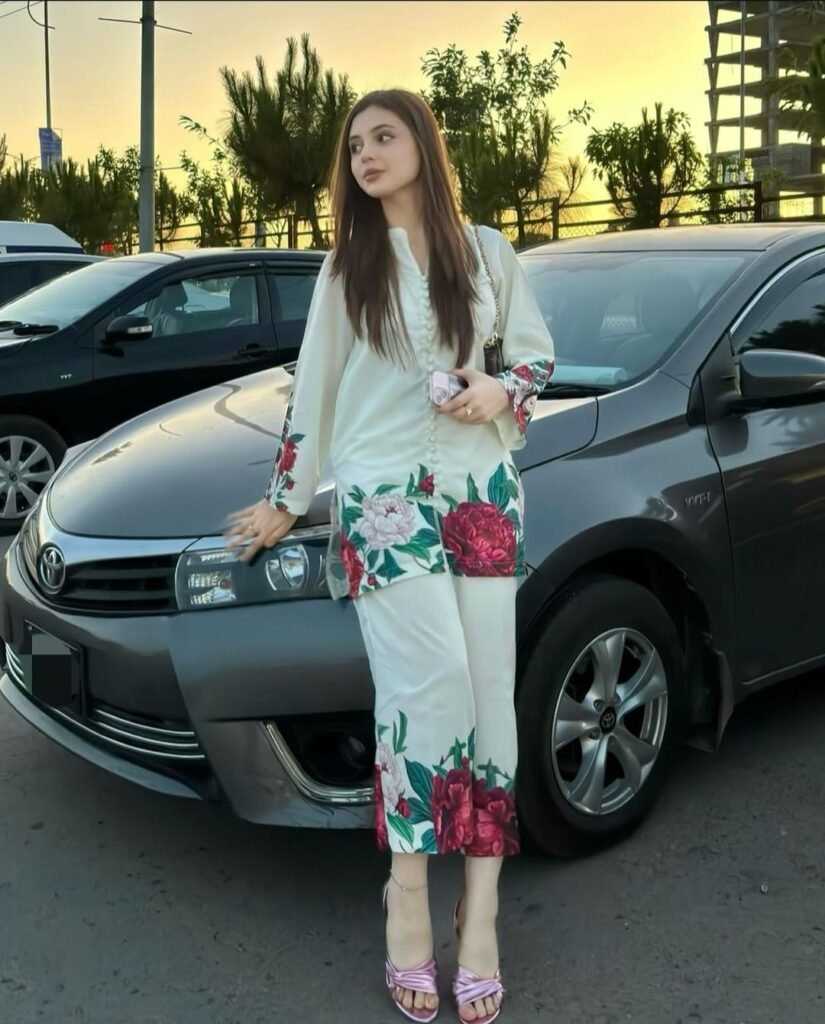 A woman with long brown hair, wearing a white outfit with pink floral patterns, stands in front of a parked grey car at sunset. She is leaning slightly on the car and looking to the side.