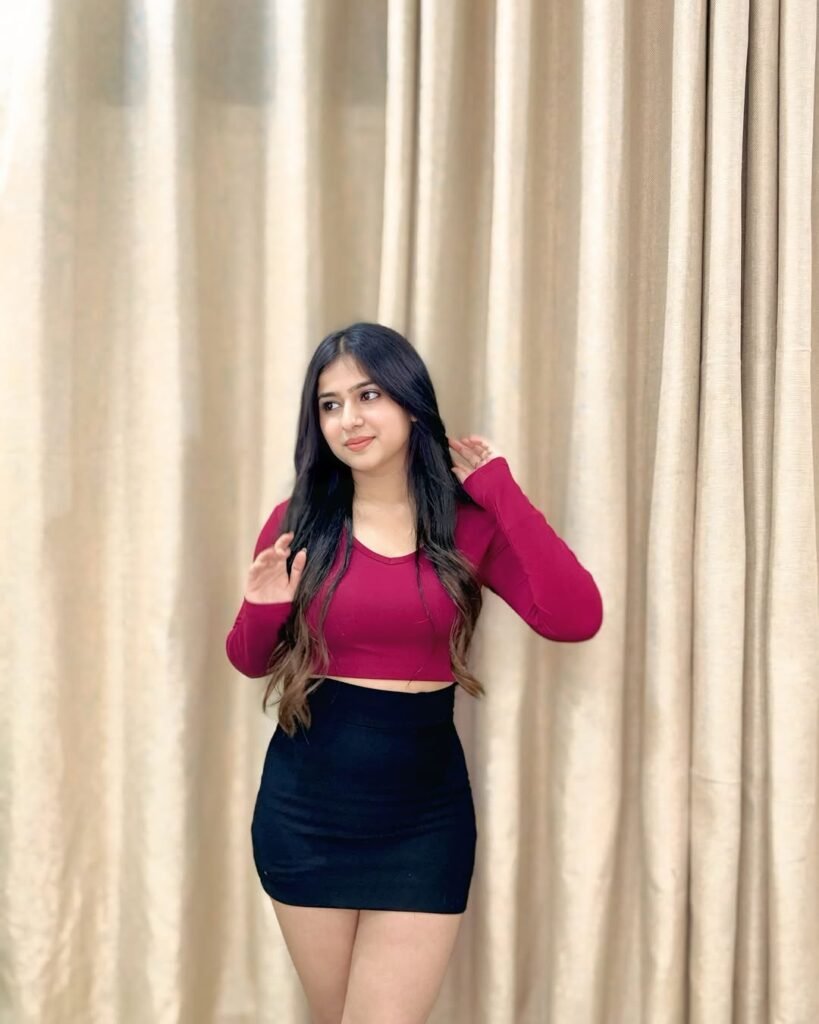 A young woman with long dark hair is standing in front of beige curtains, wearing a maroon long-sleeve crop top and a black mini skirt, looking slightly to the side and touching her hair with one hand.