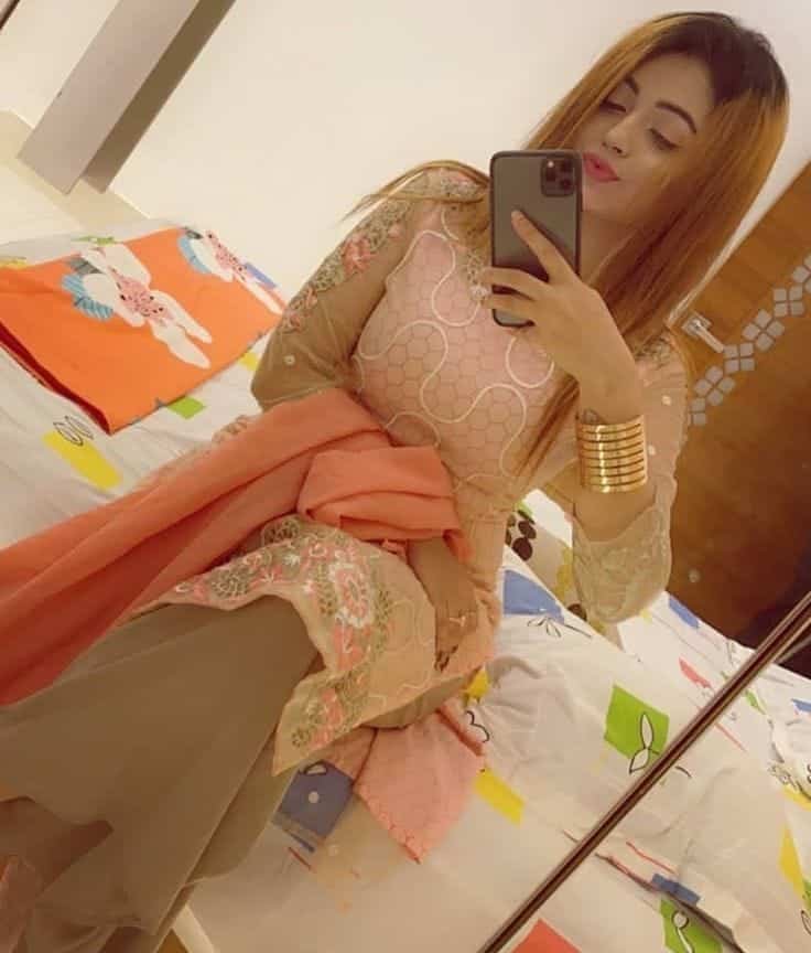 A woman in a peach and beige traditional outfit takes a mirror selfie while sitting on a bed with colorful sheets and an orange pillow, holding her phone in one hand and wearing gold bangles.