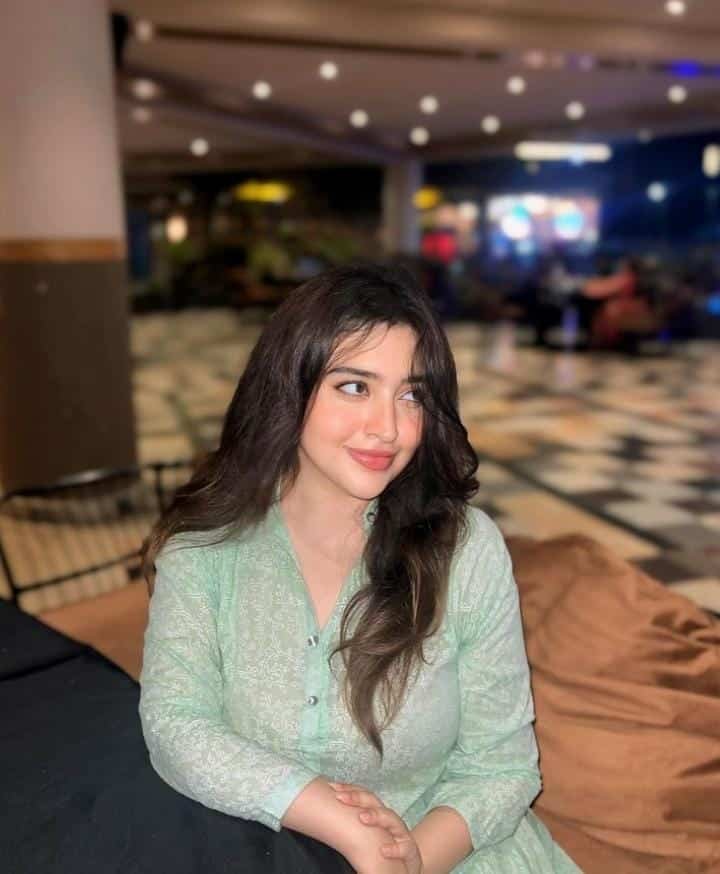 Anusha Khan in a green dress, seated on a bed, exuding a calm and relaxed demeanor.